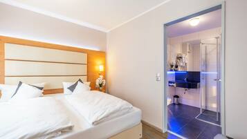 Une chambre d’hôtel avec un lit, une table de chevet, une lampe fixée au mur et une salle de bain visible à travers une porte ouverte.