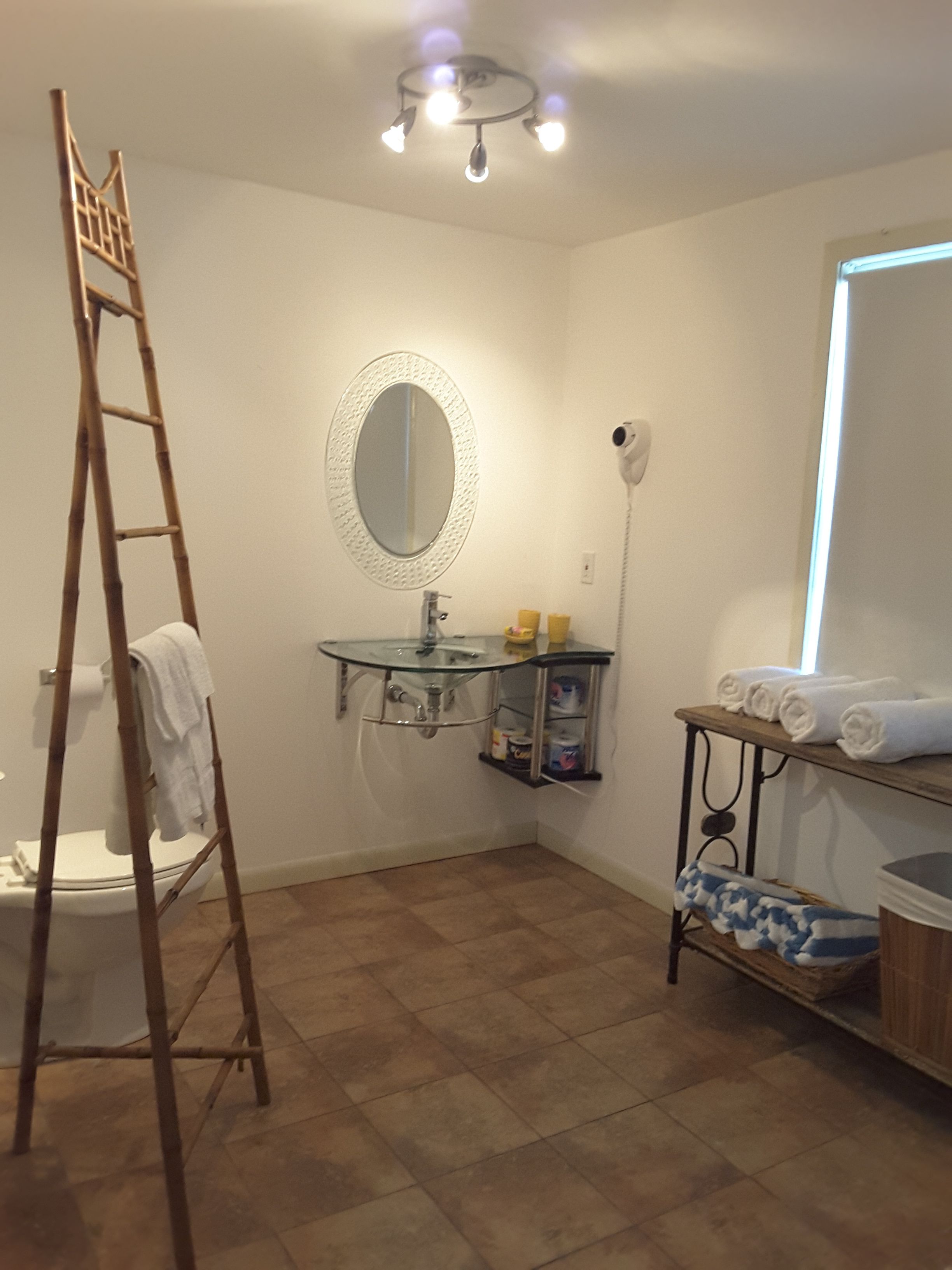 Un baño con inodoro, un lavabo con encimera de vidrio, espejo, secador de pelo, estante con toallas y una escalera.