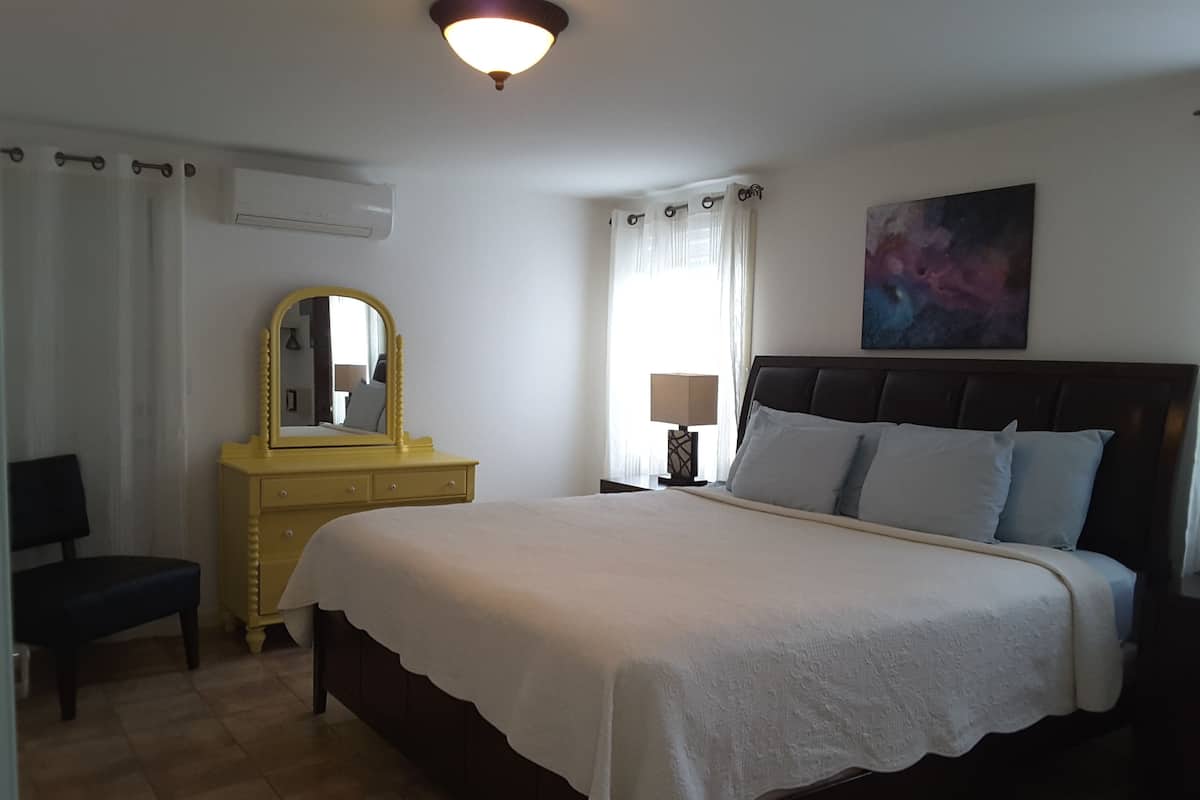 A bedroom with a large bed, a dresser, a mirror, a nightstand with a lamp, and a painting on the wall.