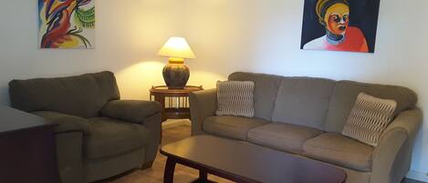 A living room with a sofa, two armchairs, a coffee table, a lamp, and two paintings on the walls.