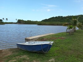 A blue boat tied to a concrete dock by a grassy shore with a bench and palm trees in the background.