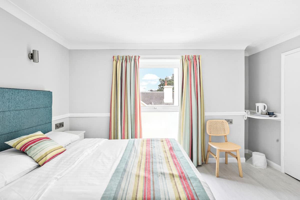 Une chambre d’hôtel moderne équipée d’un lit, d’une chaise, d’une fenêtre avec des rideaux et d’une petite table sur laquelle se trouve une bouilloire.