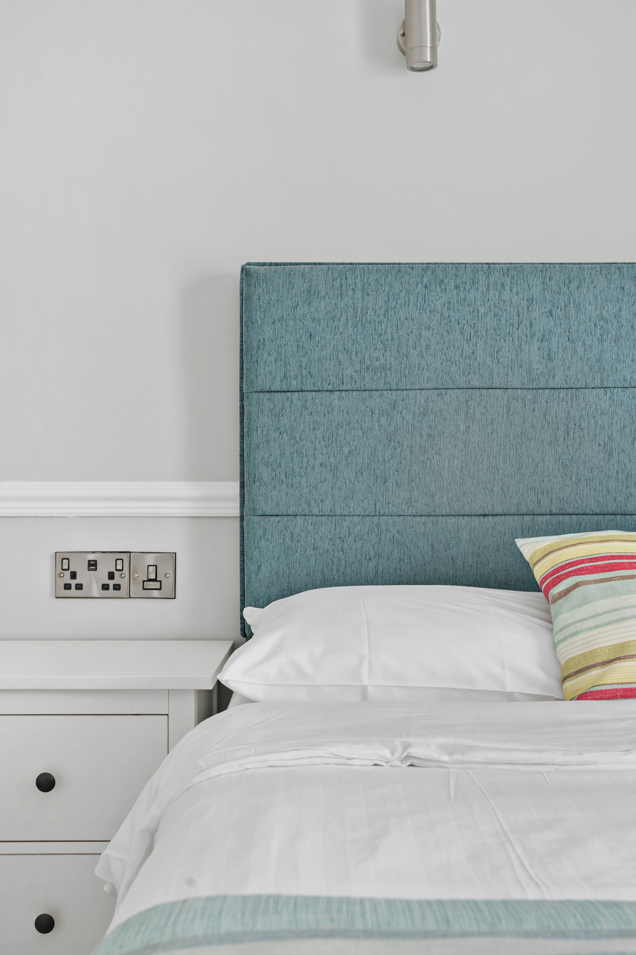 A bed with white bedding and a teal headboard, a bedside table with a power outlet, and a nightstand with drawers.