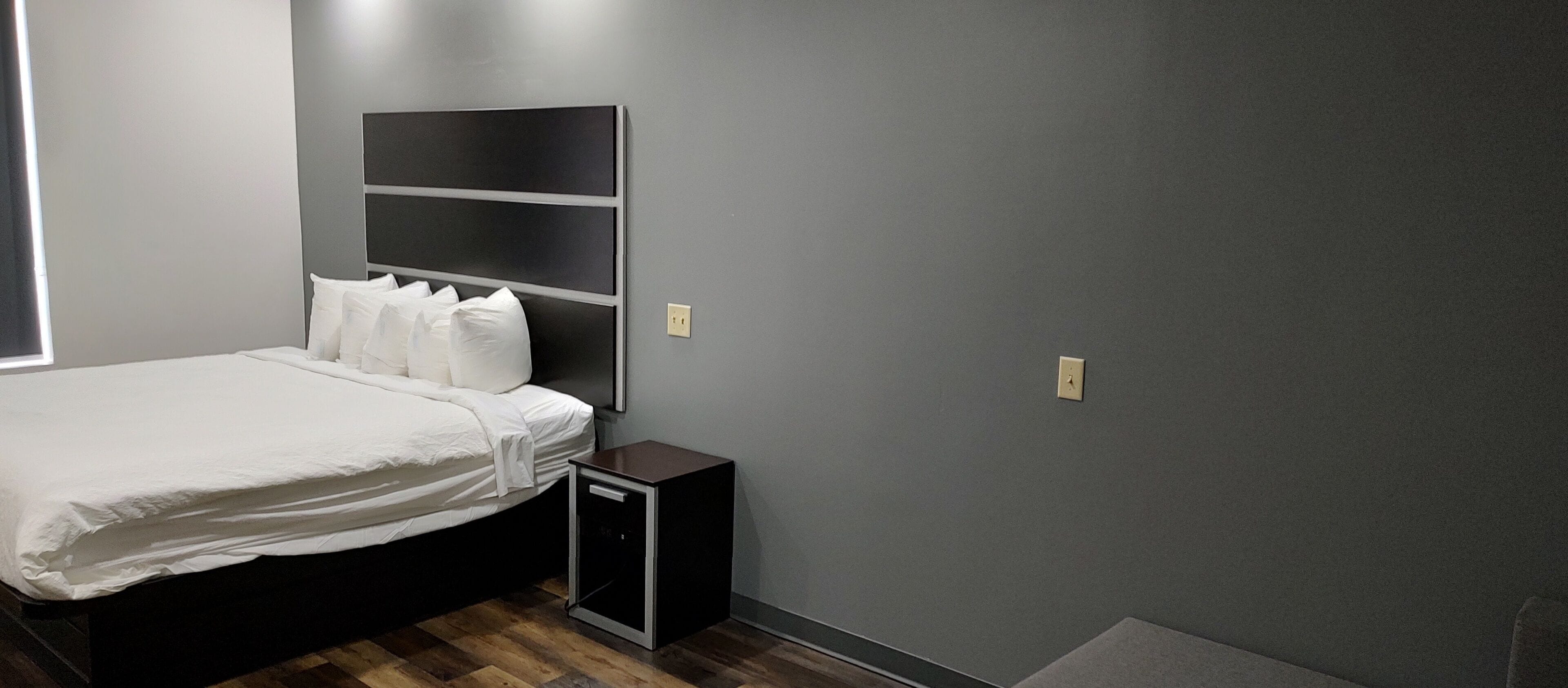 Un lit bien fait dans une pièce dont le mur est peint en gris foncé, avec une petite table de chevet et une étagère fixée au mur au-dessus du lit.