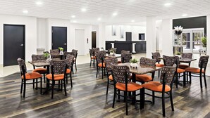 Un comedor amplio con varias mesas y sillas, una cocina moderna al fondo, y un ambiente limpio y bien iluminado.