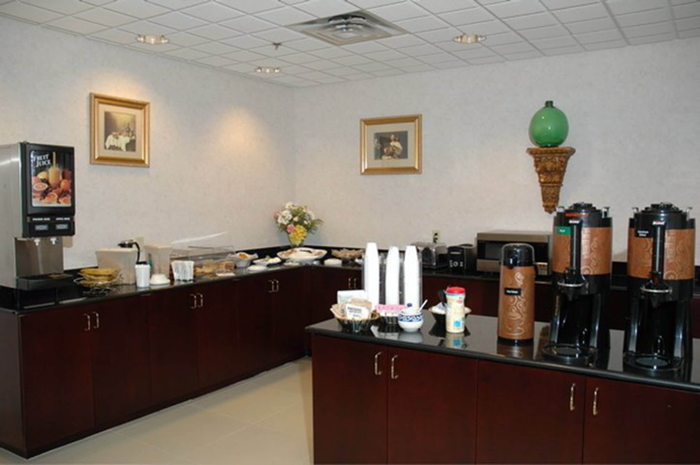 A hotel room with a kitchenette, a refrigerator, a coffee machine, and a vase with flowers on the counter.