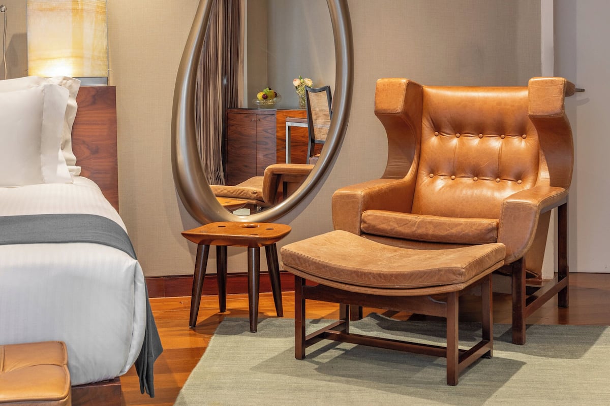 A modern hotel room with a large mirror, a leather armchair, a wooden side table, and a bed with a white comforter.