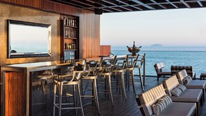 Zona de bar con vistas al mar, terraza de madera, taburetes y sillones de descanso.