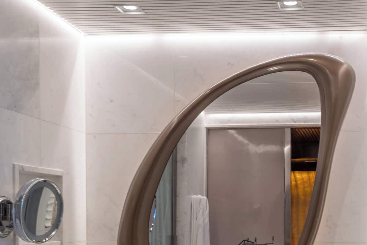A modern bathroom with a large oval mirror, a white sink, and a glass shelf with toiletries.
