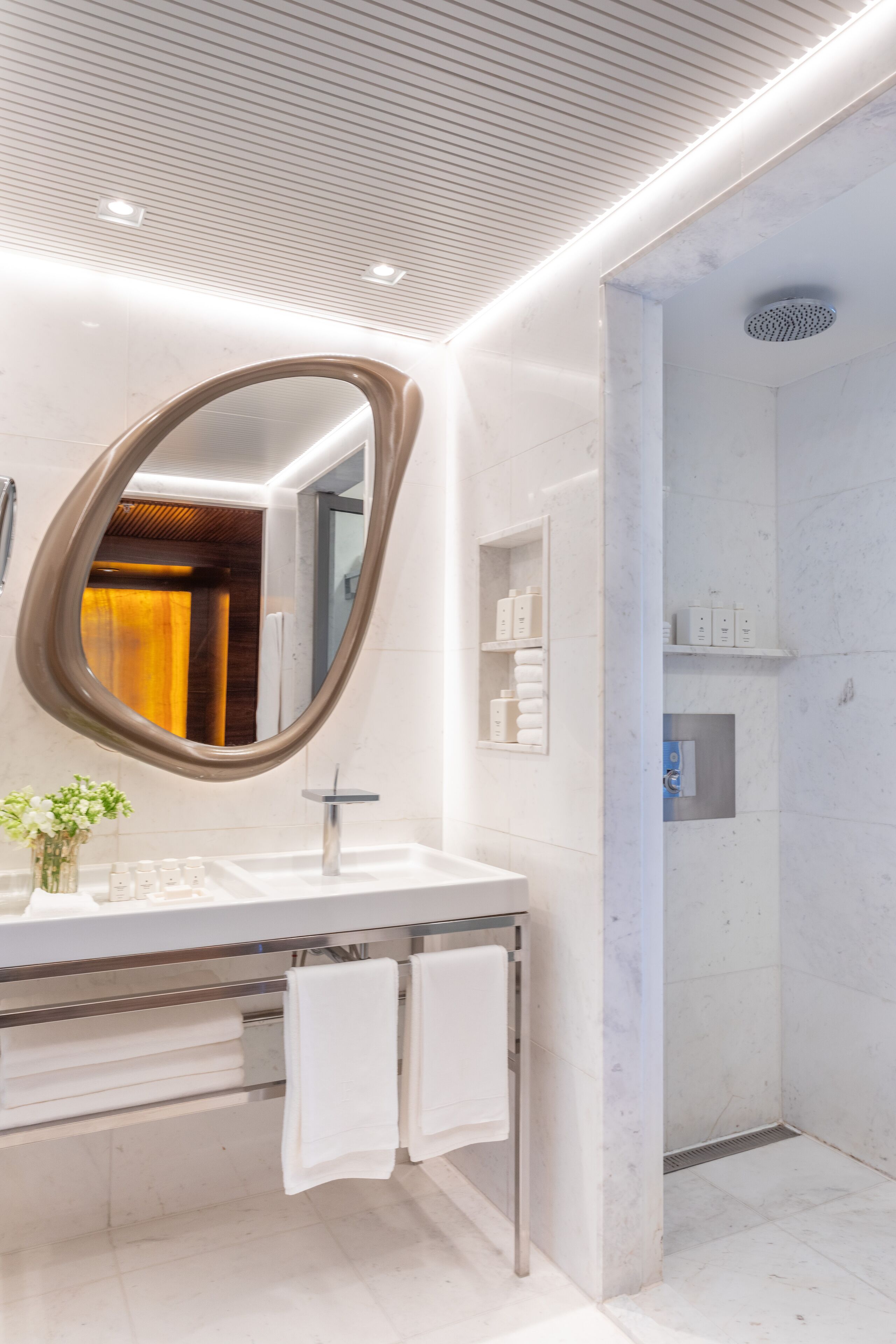 Een moderne badkamer met een grote, ovale spiegel, witte marmeren tegels, een glazen wastafelmeubel en een douchegedeelte.