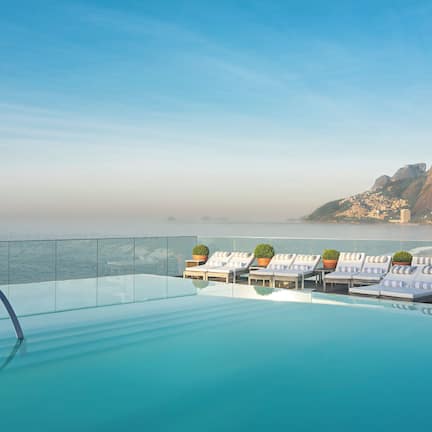 A rooftop infinity pool with lounge chairs and potted plants, overlooking a beach and mountains.