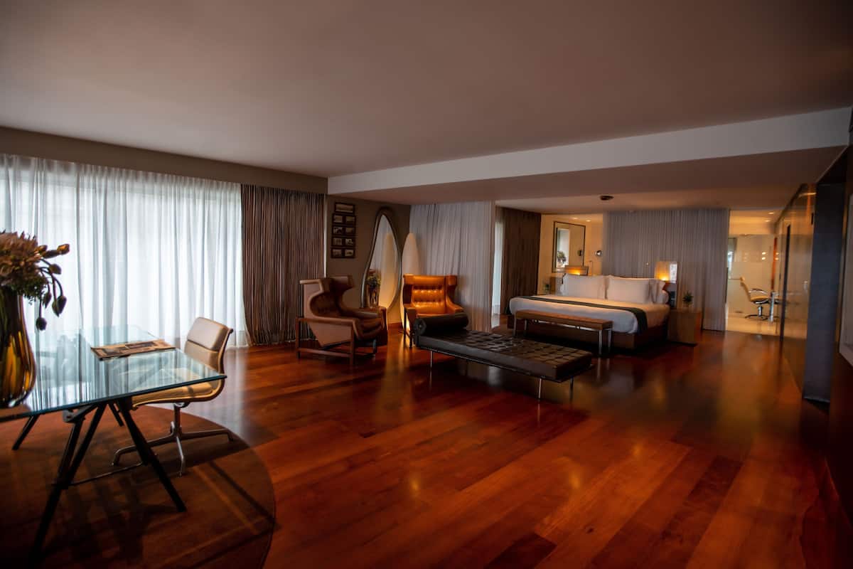 A modern hotel room with a glass dining table, leather chairs, a sofa, a bed, and a large mirror.