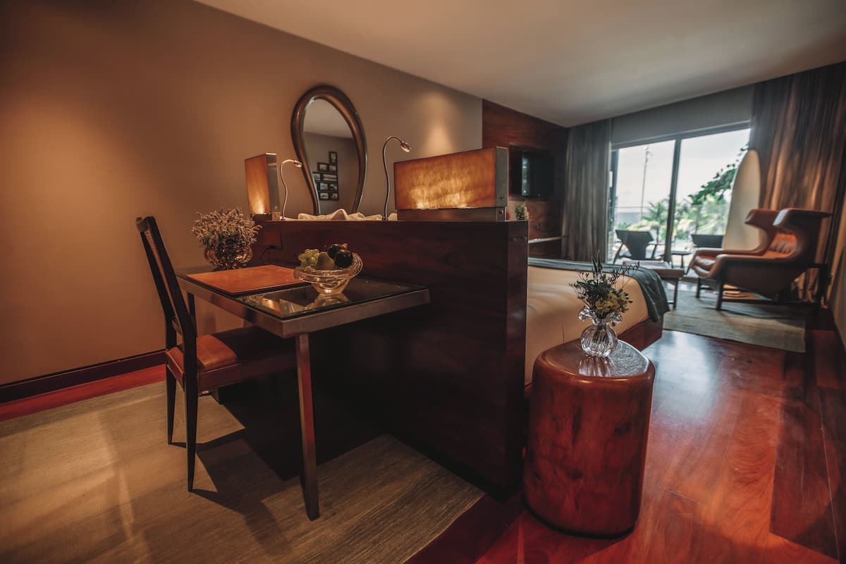 A modern hotel room with a wooden dining table, a chair, a small round table with a vase, and a large window offering a view of the outdoors.