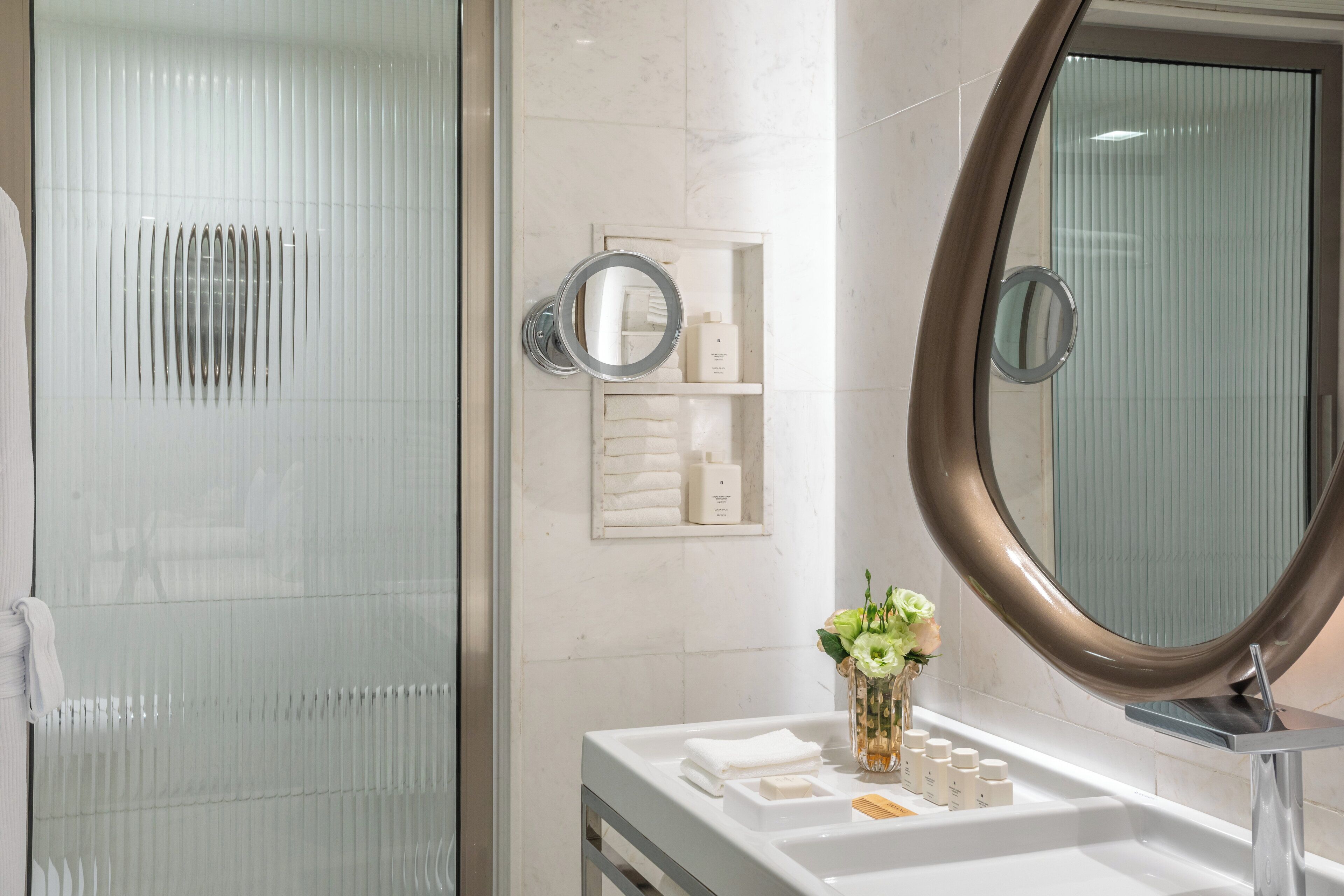 A modern bathroom with a glass shower enclosure, a vanity with a large mirror, and a small shelf with toiletries.