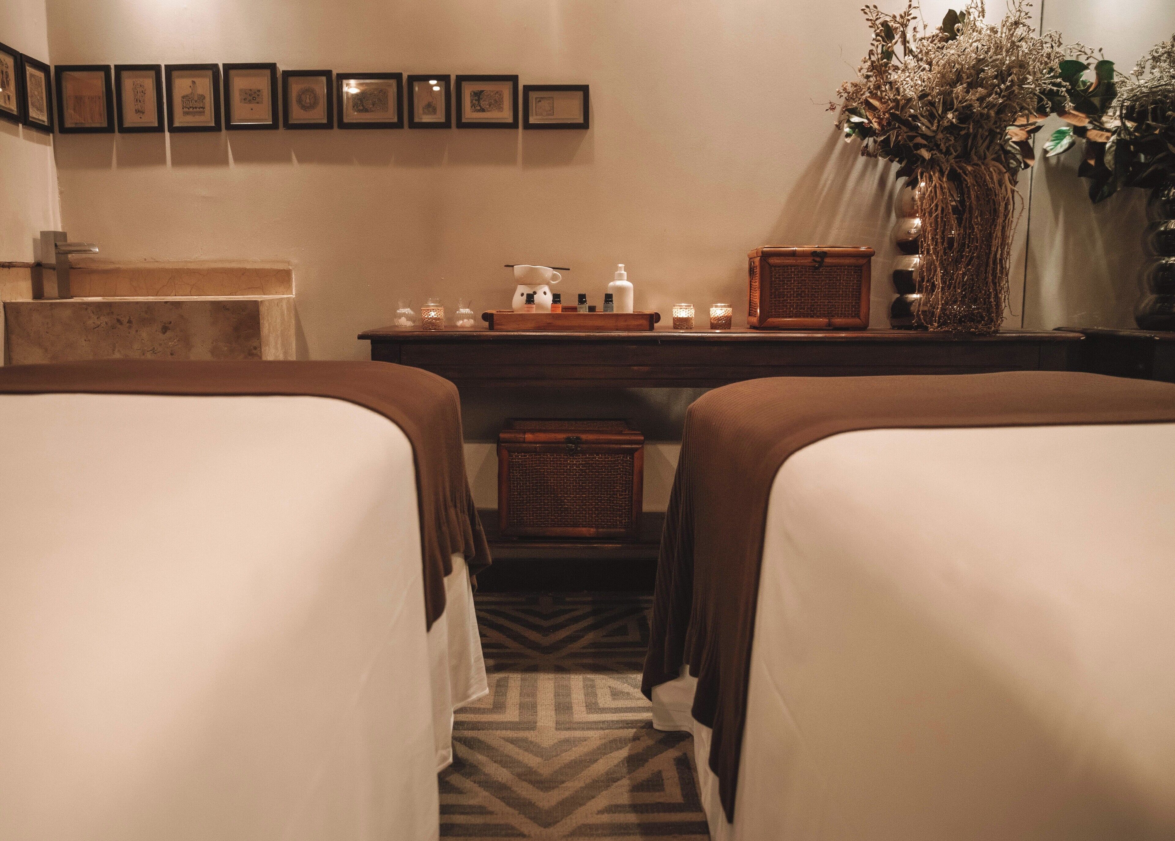 A hotel room with two beds, a wooden desk with toiletries, framed pictures on the wall, and a potted plant.