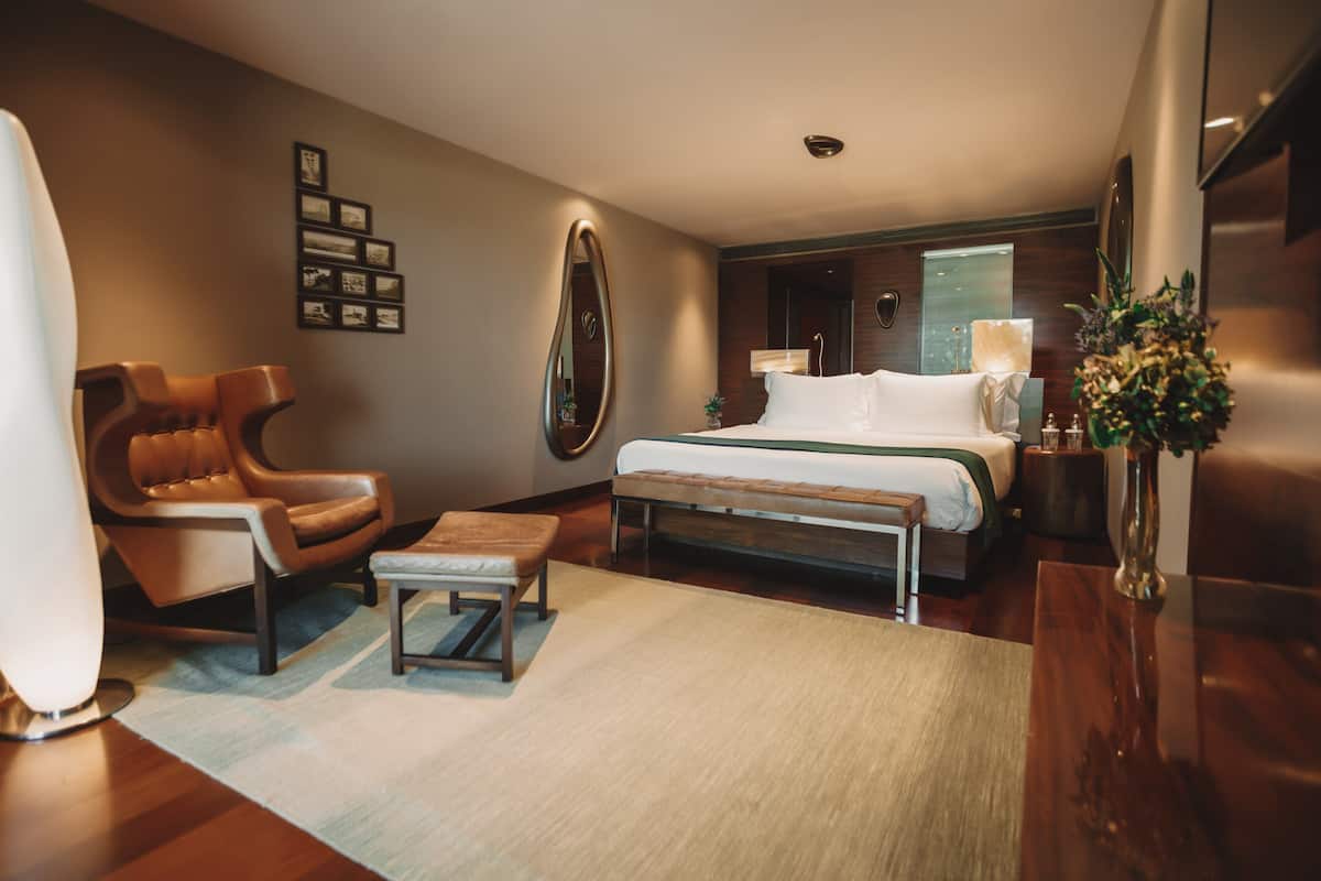 A modern hotel room with a bed, a leather armchair, a small table, and a vase with flowers.