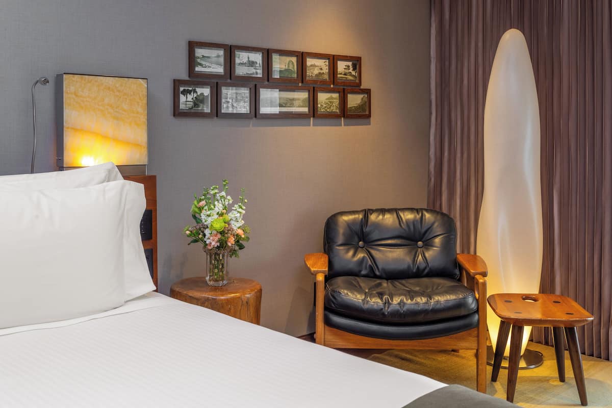 A hotel room with a bed, a black leather chair, a wooden side table with a vase of flowers, a wall-mounted picture frame, and a large window with curtains.