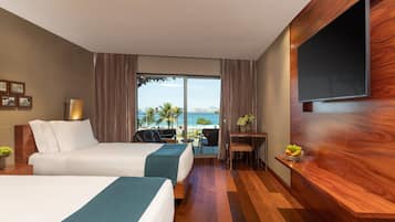 A hotel room with two beds, a TV, a desk, and a view of the ocean.
