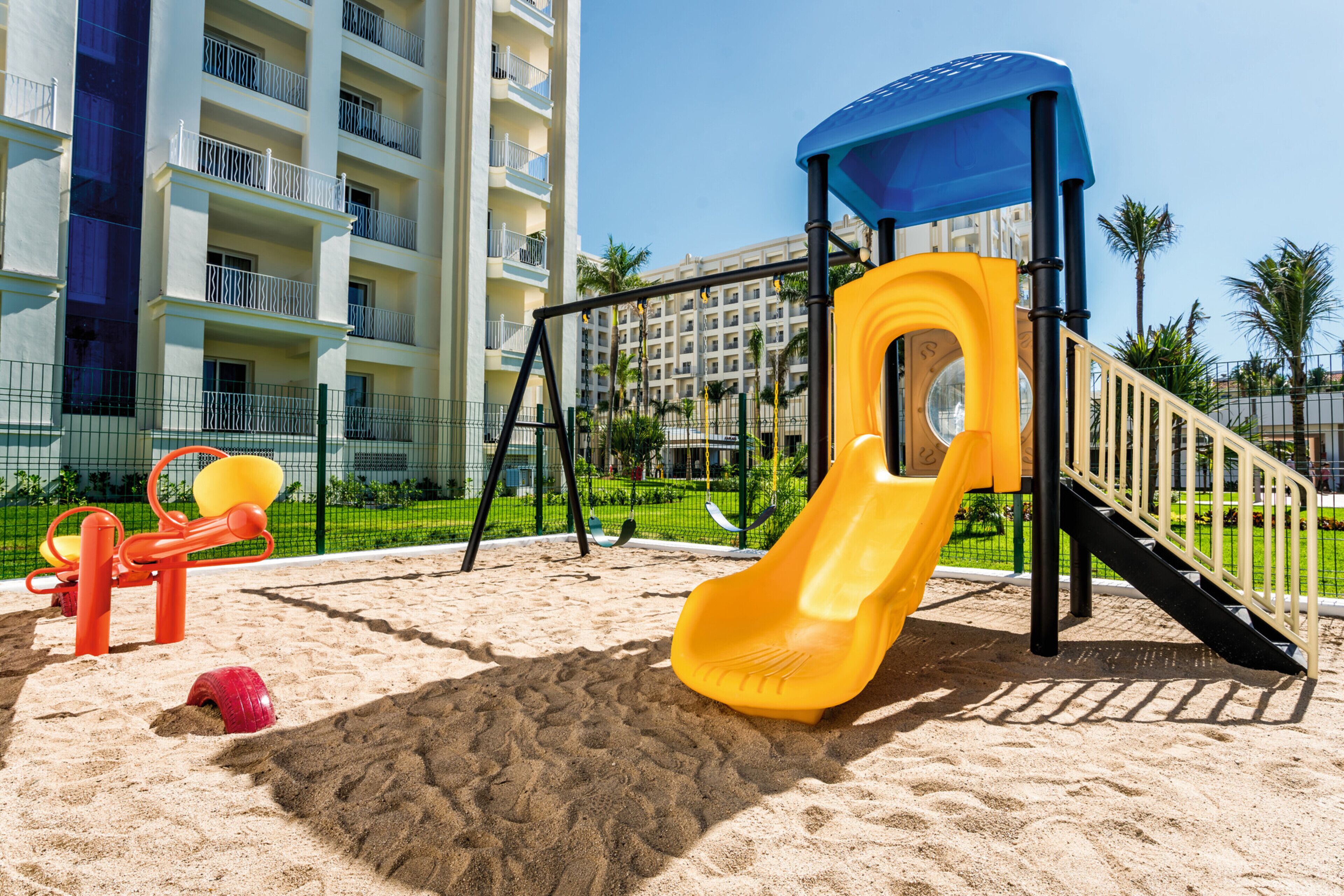 Un parque infantil con un tobogán amarillo y un balancín rojo, rodeado de arena y una valla.