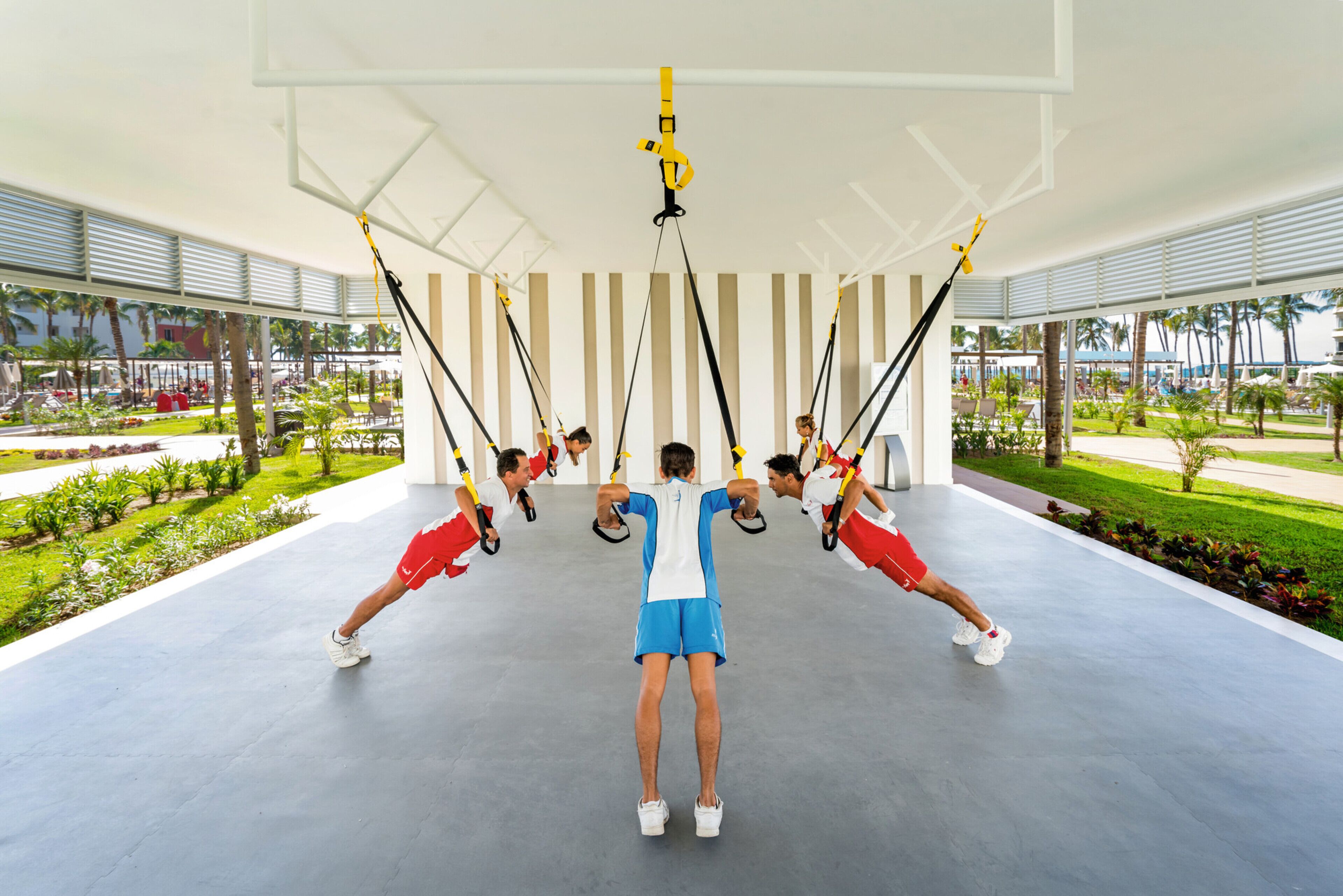 A group of people doing TRX exercises in a covered outdoor area with a view of a landscaped garden and palm trees.