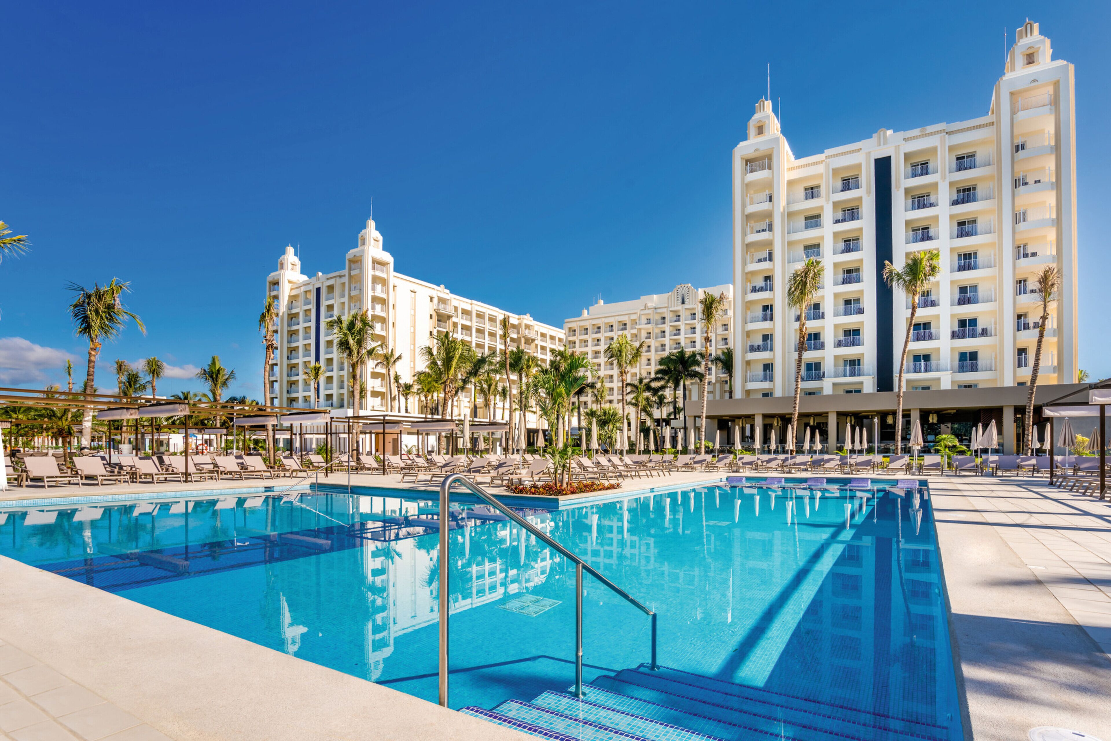 A hotel pool area with multiple lounge chairs and umbrellas, surrounded by palm trees and a large building with multiple balconies.