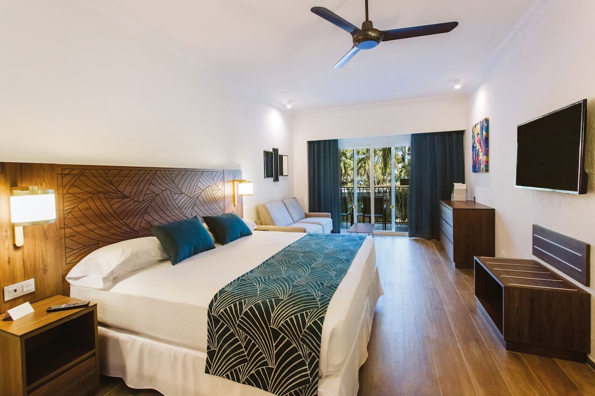 Habitación de hotel moderna con una cama grande, ventilador de techo, televisión y balcón con vista a palmeras.