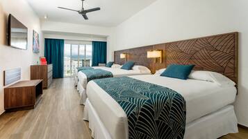 Habitación de hotel con dos camas, un escritorio, un ventilador de techo y un ventanal grande con cortinas.
