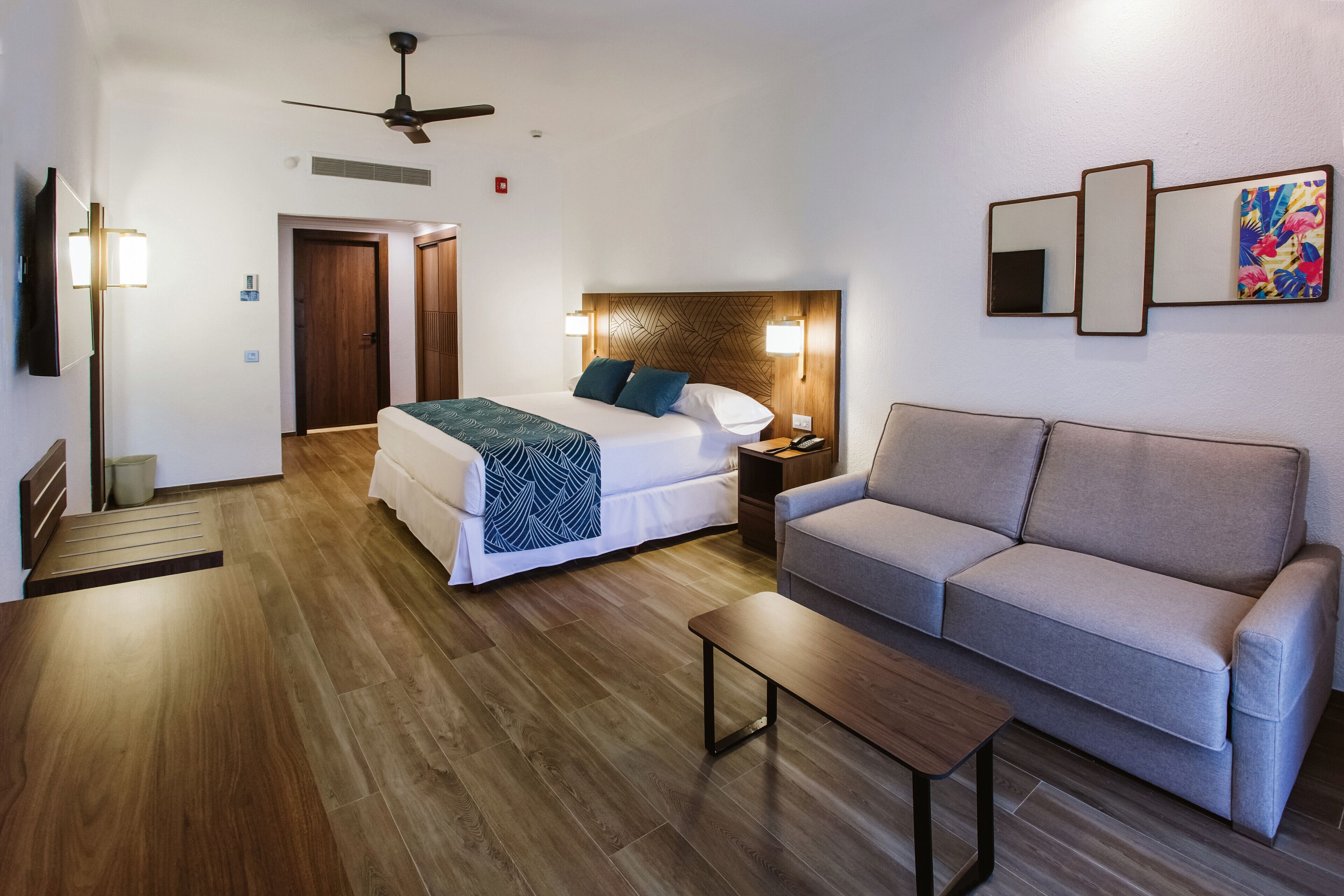 Una habitación de hotel con una cama, un sofá, una mesa de centro, un ventilador de techo y un televisor montado en la pared.