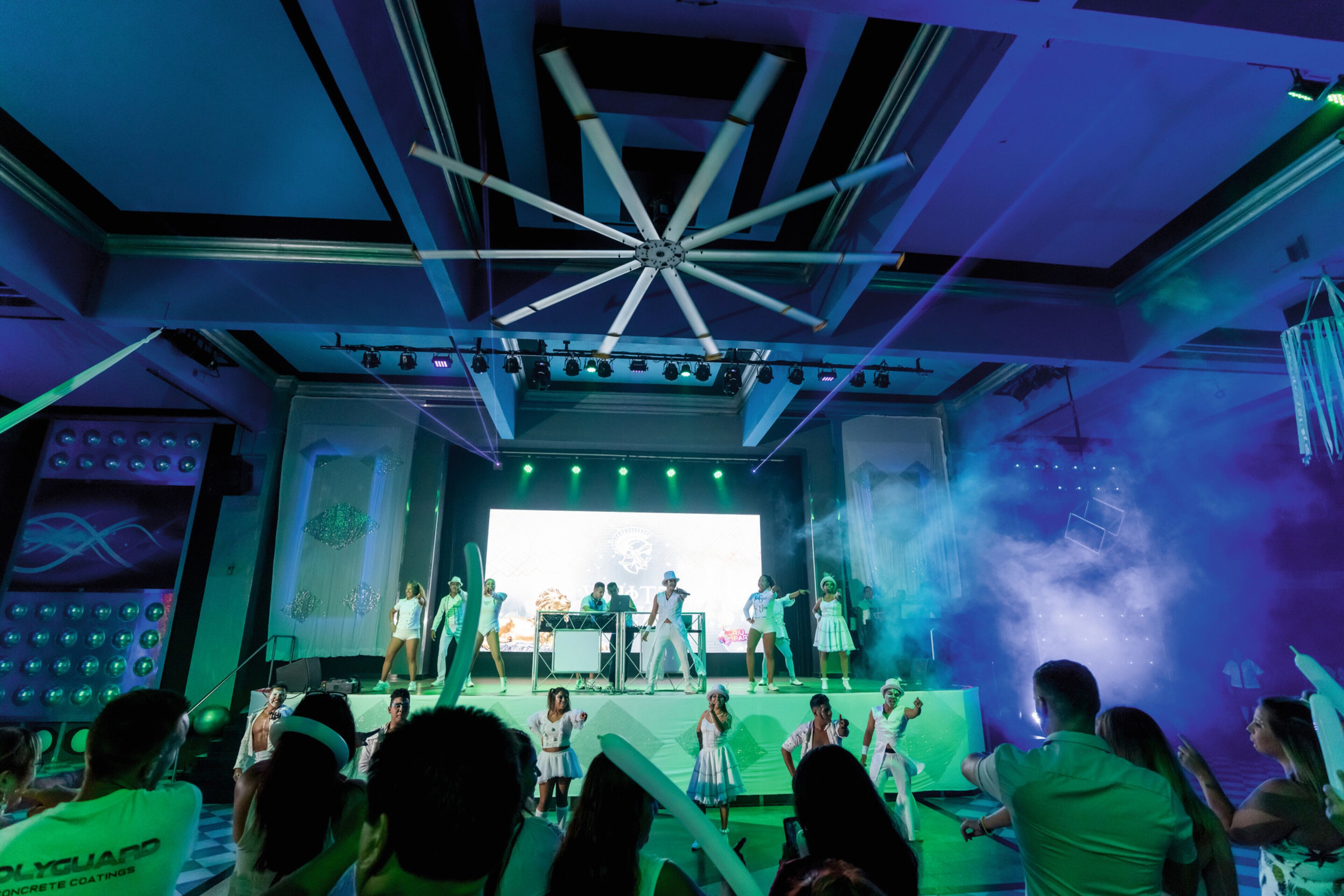 Un escenario con artistas y público en un recinto con diseño de techo moderno y una pantalla grande al fondo.