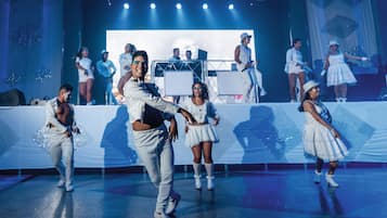 Palco com artistas em trajes coordenados, uma tela grande ao fundo e iluminação cênica.