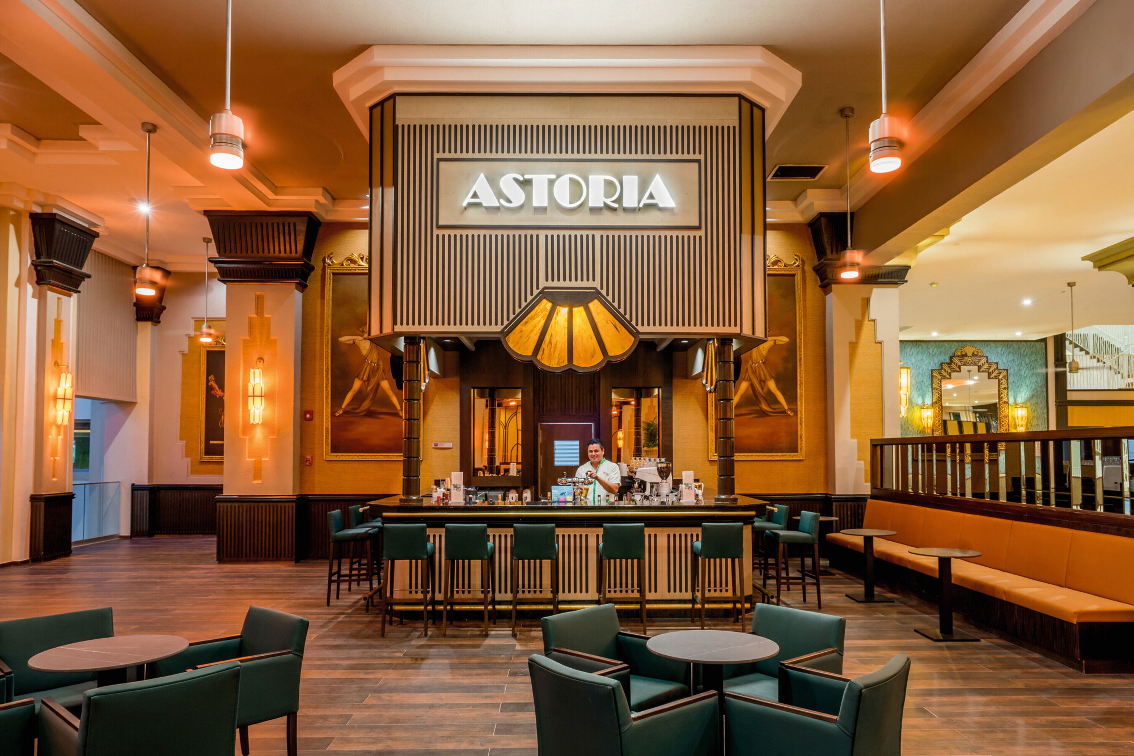 Amplia zona de bar con un llamativo letrero 'ASTORIA', diversas opciones de asientos y una barra.