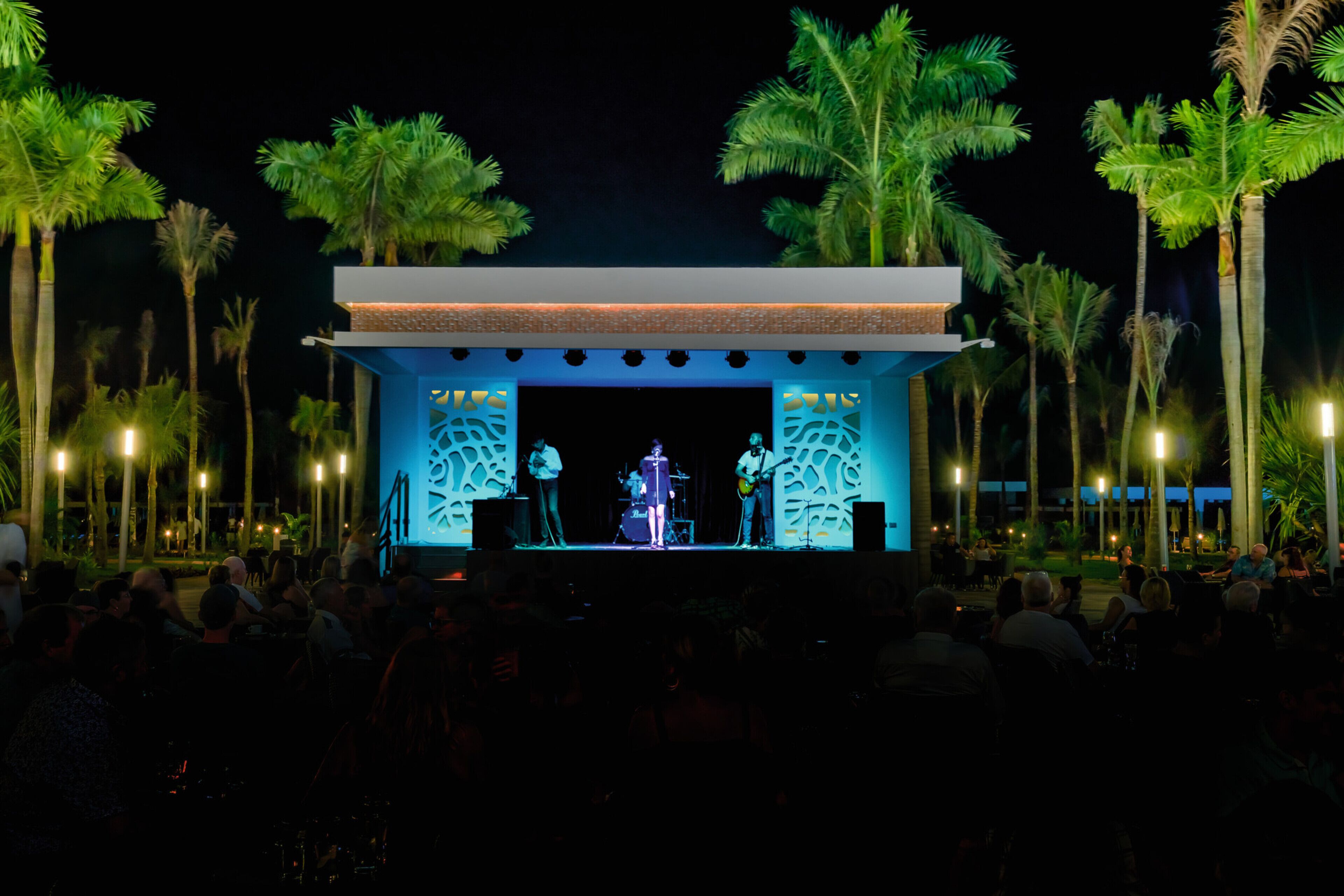 An outdoor stage with a band performing, surrounded by palm trees and a seated audience.