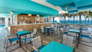 A modern restaurant with a wooden bar and teal-colored furniture, overlooking a beach. - Hotel Riu Vallarta - All Inclusive (Nuevo Nayarit)