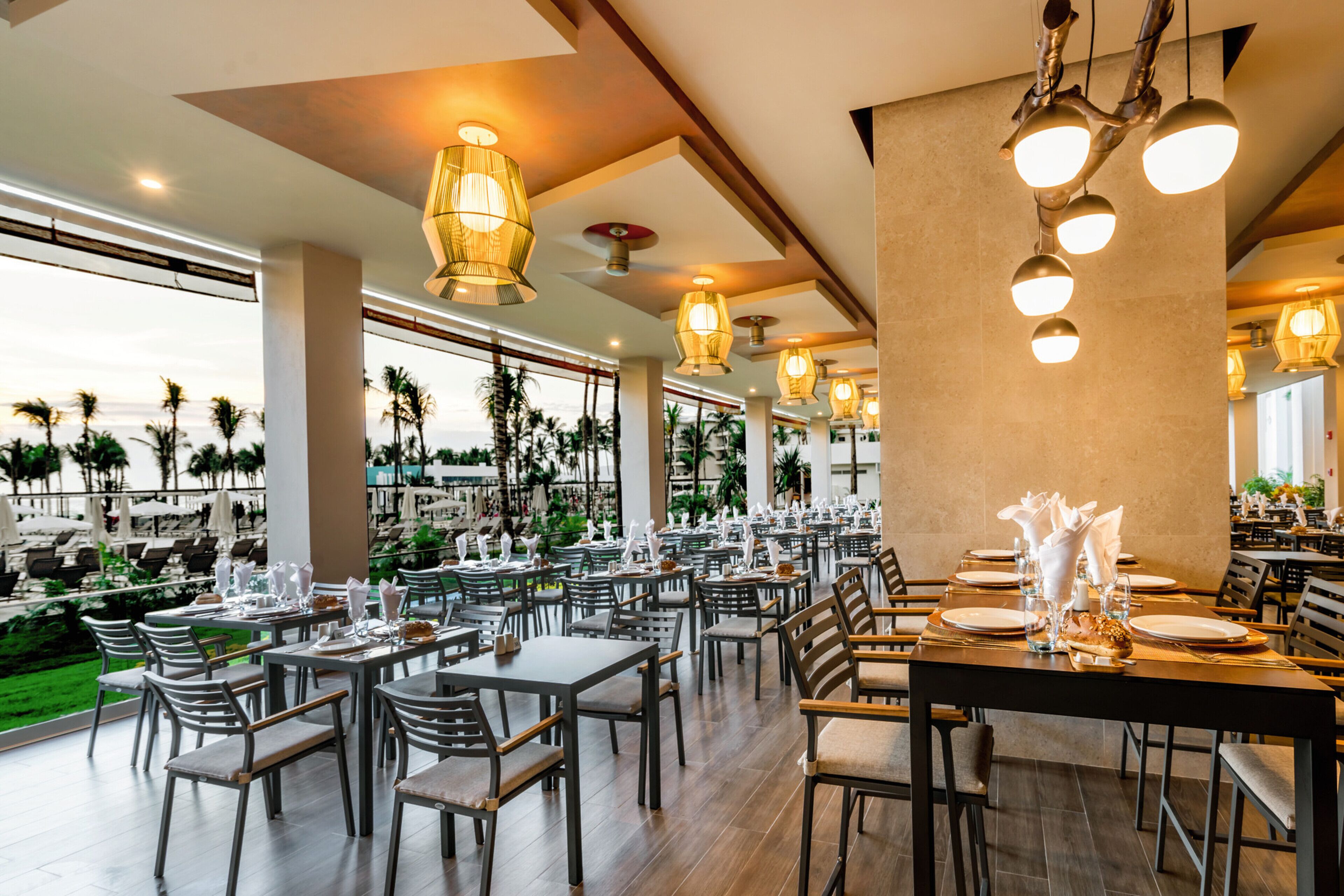 A restaurant with multiple dining tables and chairs, large windows offering a view of palm trees and the ocean, and hanging pendant lights.