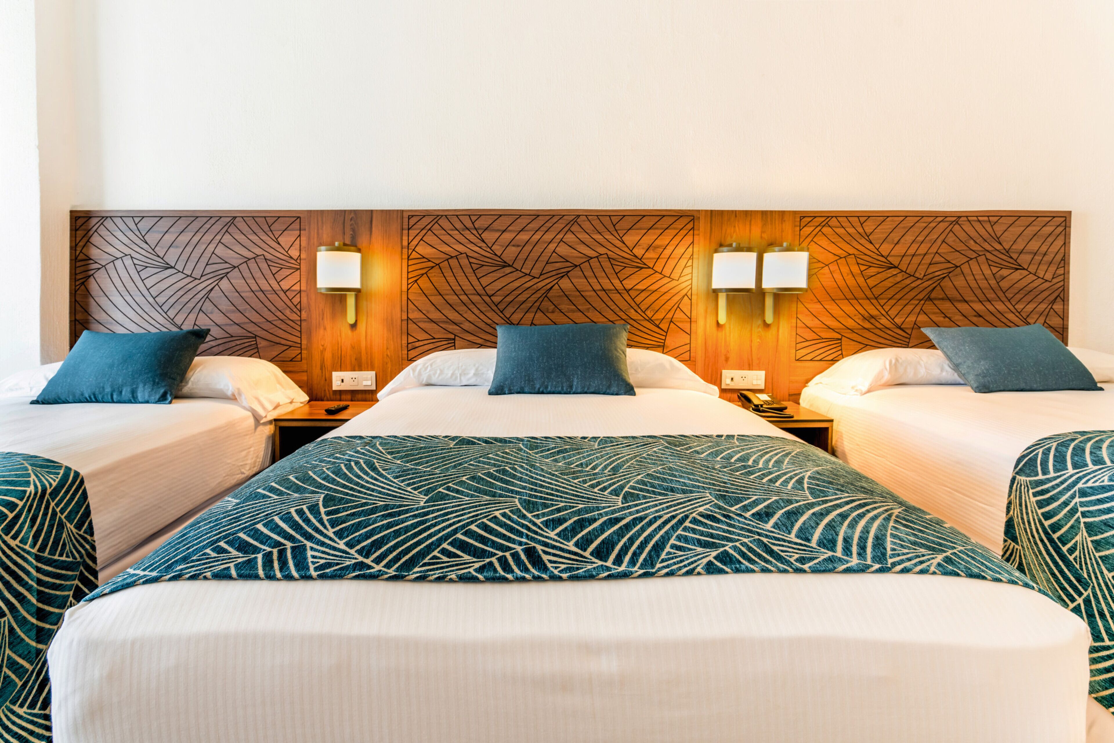 A hotel room with three beds, each with a blue pillow and a patterned bedspread. There are wall-mounted lamps on either side of the beds.