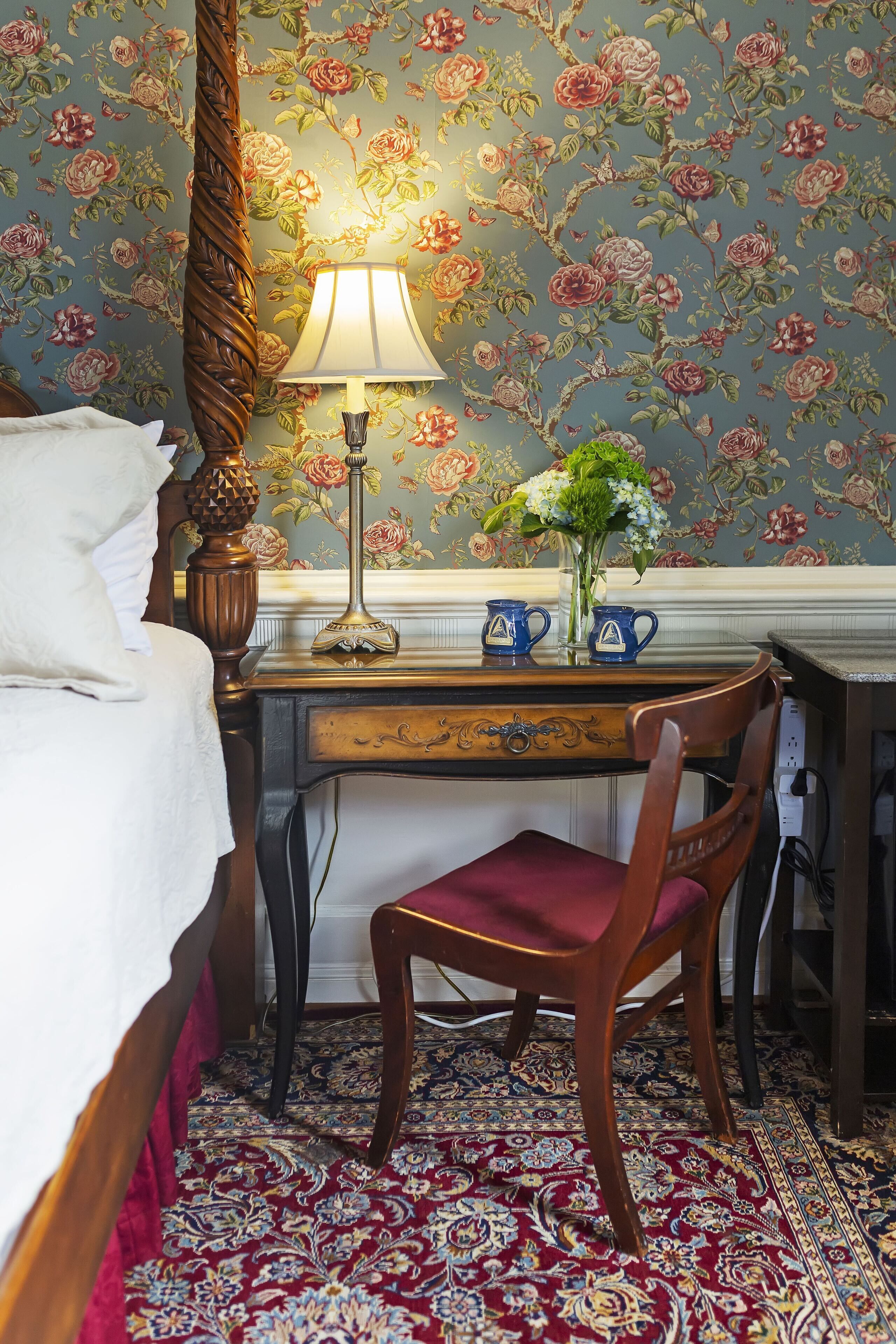 A bedroom with a bed, a desk with a lamp, a chair, and a floral wallpaper.