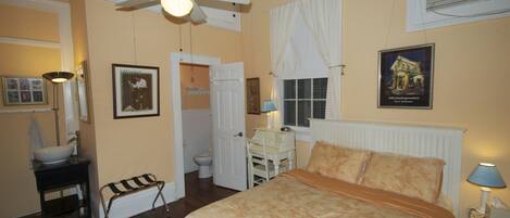 A bedroom with a bed, a ceiling fan, a window with curtains, a bedside table with a lamp, and a bathroom visible through an open door.