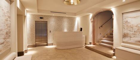 Een moderne hotel lobby met een marmeren vloer, een centrale zithoek, een lift en een trap.