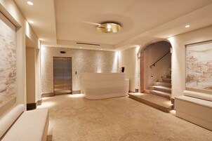 A modern hotel lobby with a marble floor, a central seating area, an elevator, and a staircase.