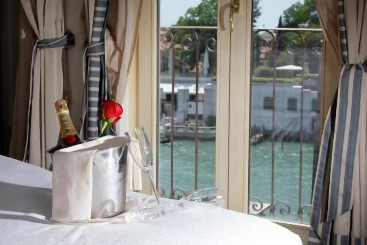 A hotel room with a bed, a champagne bottle, a glass, and a rose on the windowsill.