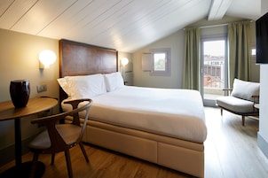 Habitación de hotel con una cama grande, un escritorio con silla, una mesita y una ventana con cortinas.