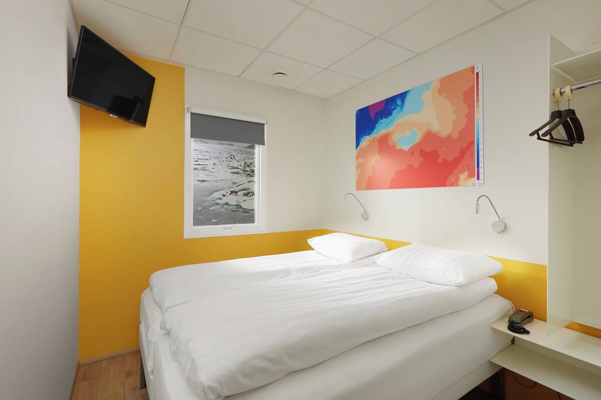 A hotel room with a bed, a television mounted on the wall, a window with a view of a snowy landscape, and a colorful abstract painting on the wall.
