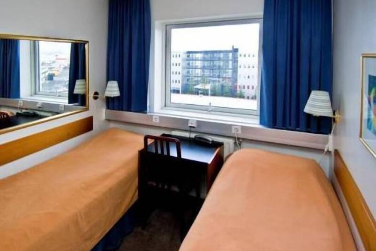 A hotel room with two single beds, a desk, a chair, a window with curtains, and a mirror.