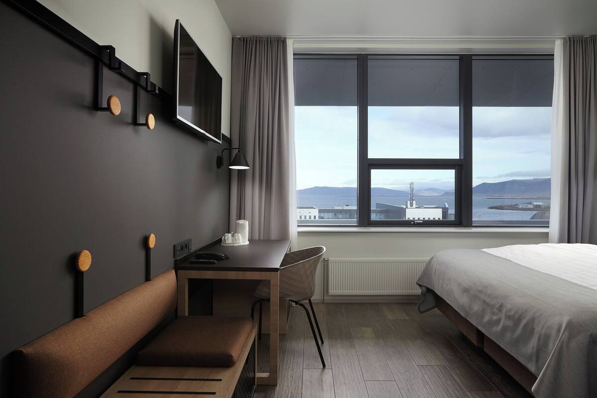 A modern hotel room with a large window, a bed, a desk, and a chair.