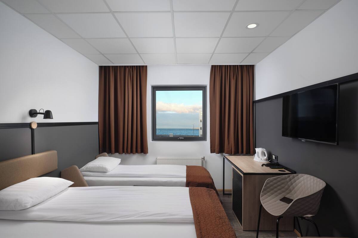 A hotel room with two beds, a desk, a chair, a TV, and a window with curtains.