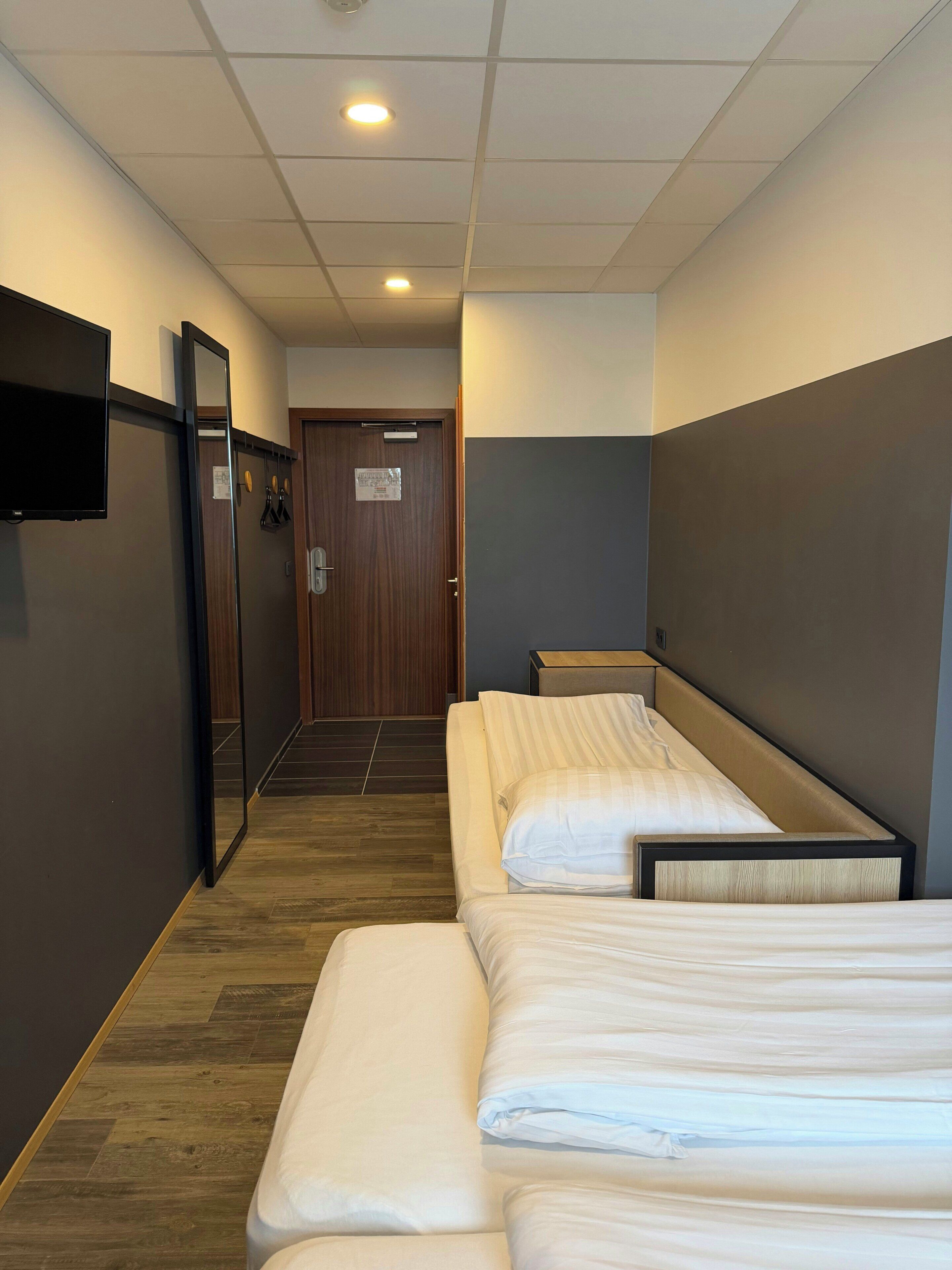 A hotel room with two single beds, a flat-screen TV mounted on the wall, a wooden door, and a tiled floor.
