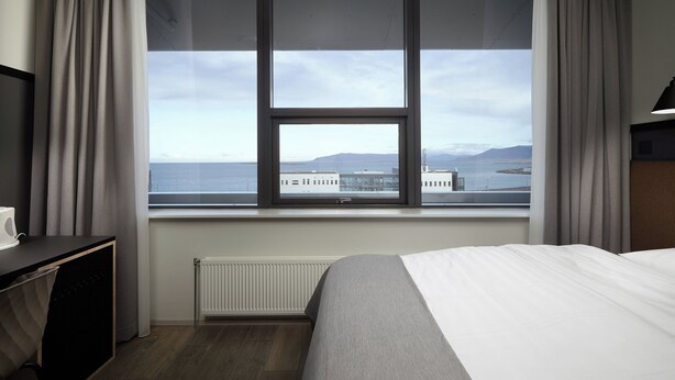 A hotel room with a large window, a bed, a desk, and a radiator. - Hotel Cabin (Reykjavik)