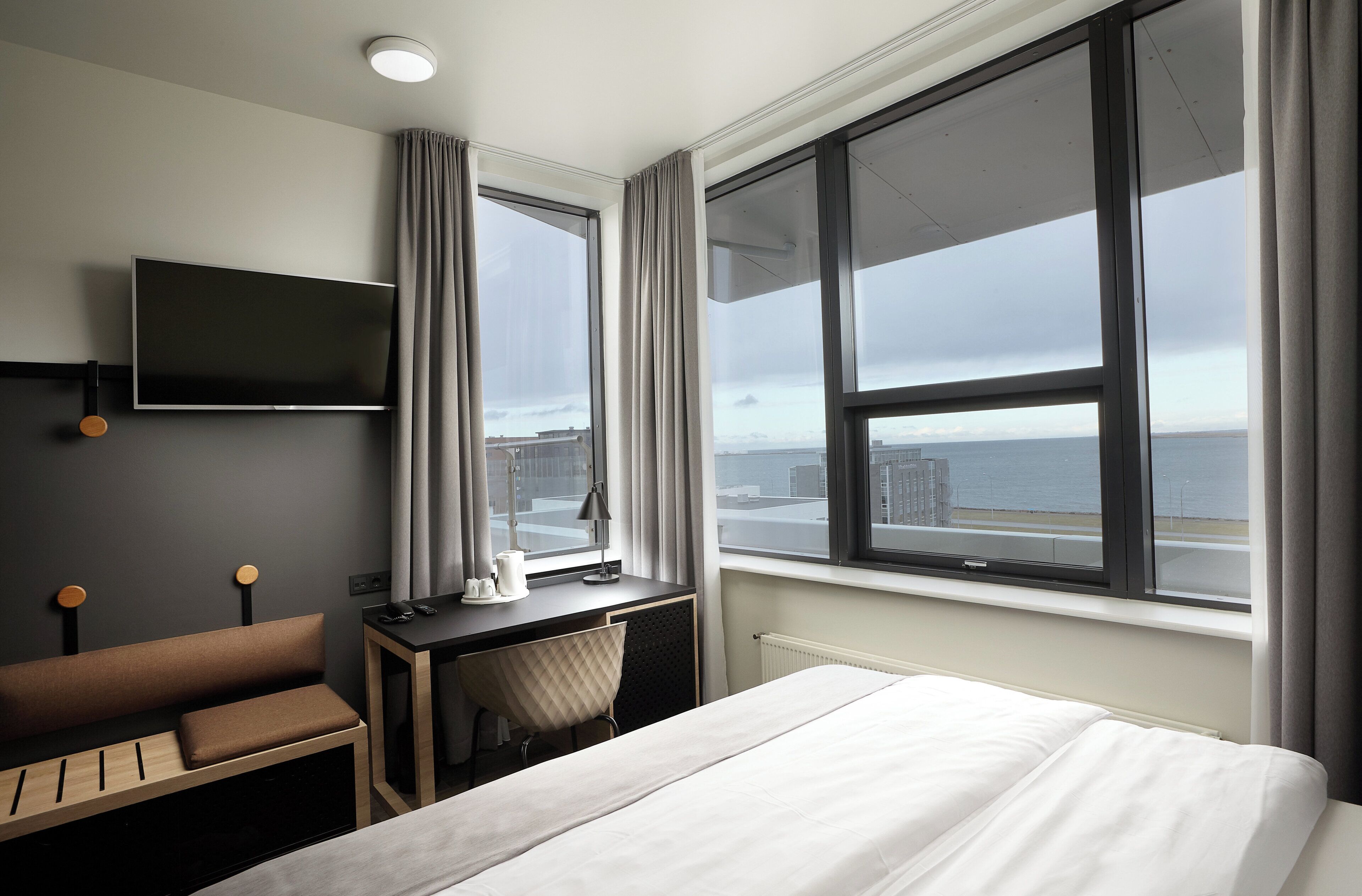Ein Hotelzimmer mit einem Bett, einem Schreibtisch mit Stuhl, einem an der Wand befestigten Fernseher und einem großen Fenster mit Blick auf Stadt und Meer.