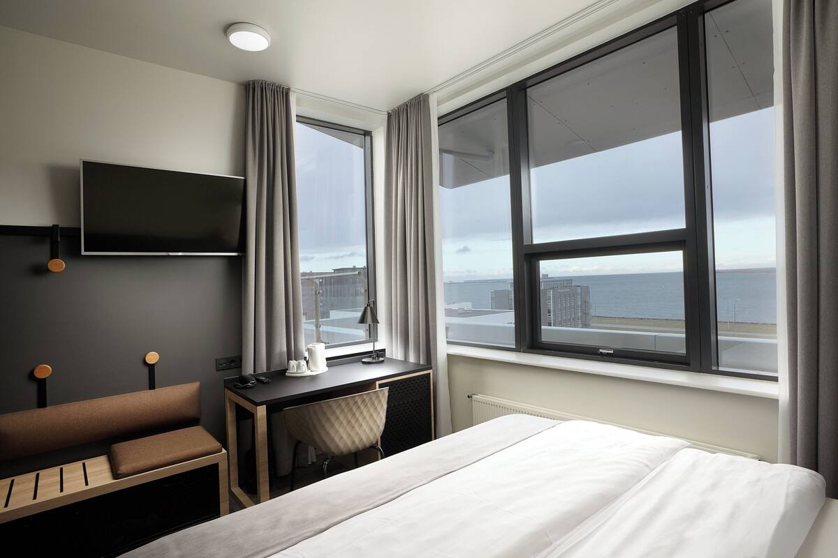 A hotel room with a bed, a desk with a chair, a TV mounted on the wall, and a large window offering a view of the city and the sea.