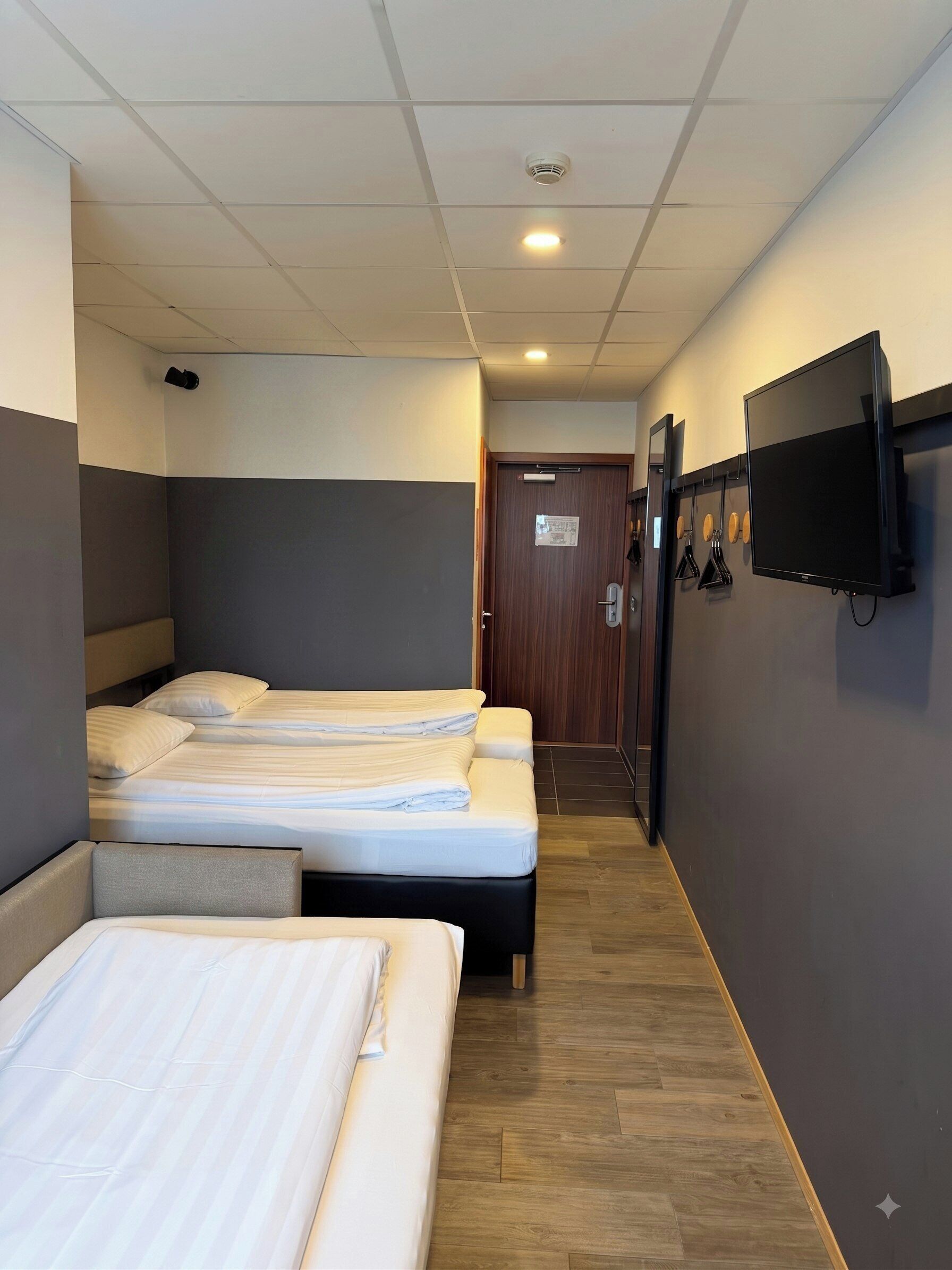 Quarto de hotel com três camas, televisão fixada na parede e chão de madeira.